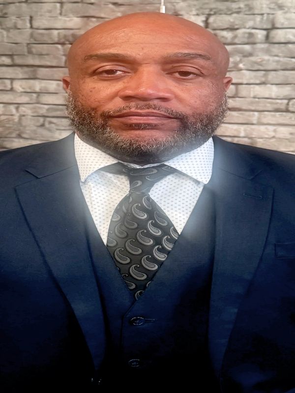 A man in a blue suit standing against a brick wall background.