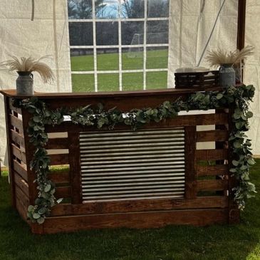 Rustic wooden bar decorated with greenery and floral arrangements under a tent.