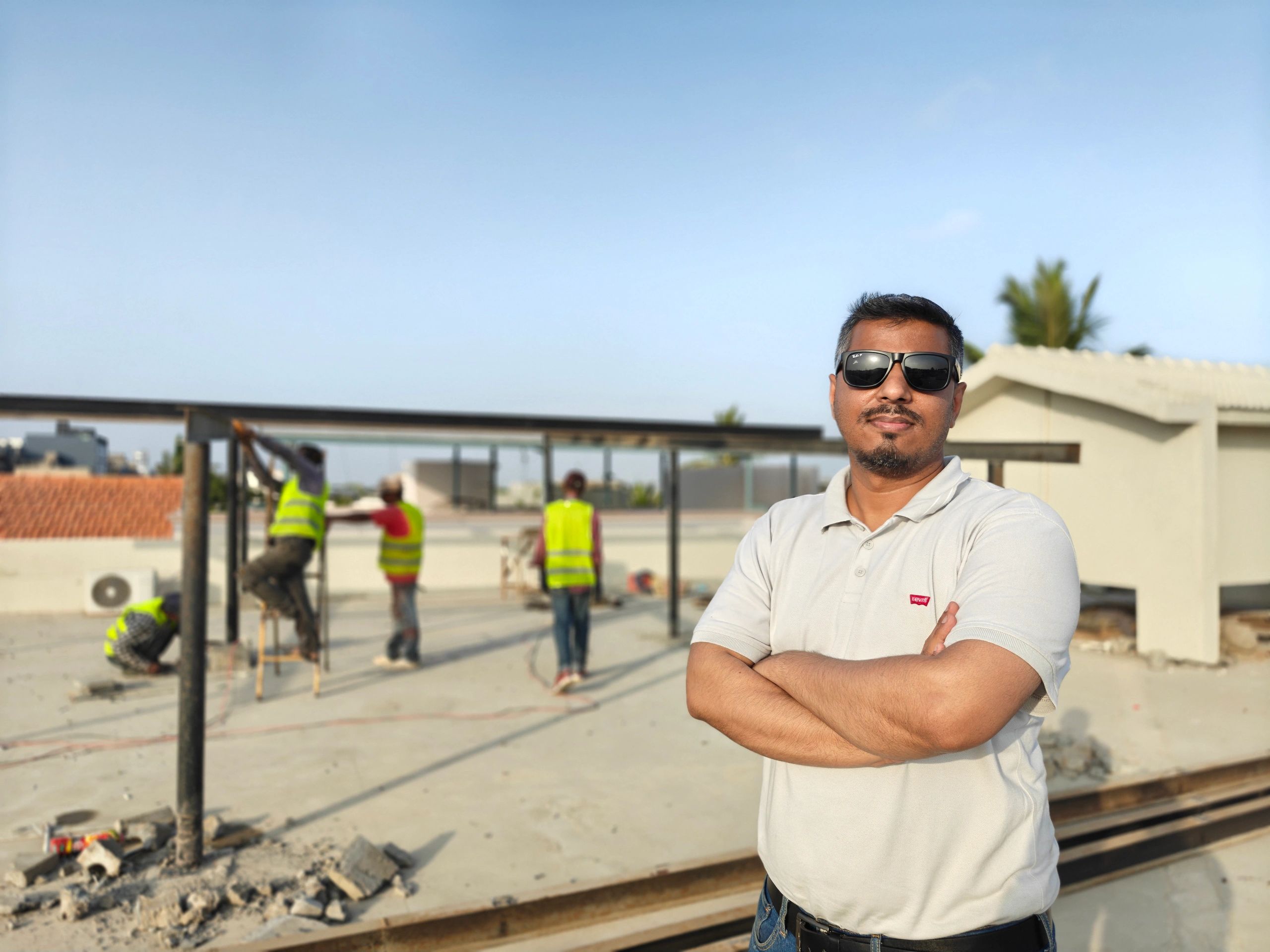 Man wearing sunglasses standing with arms crossed in front of workers.