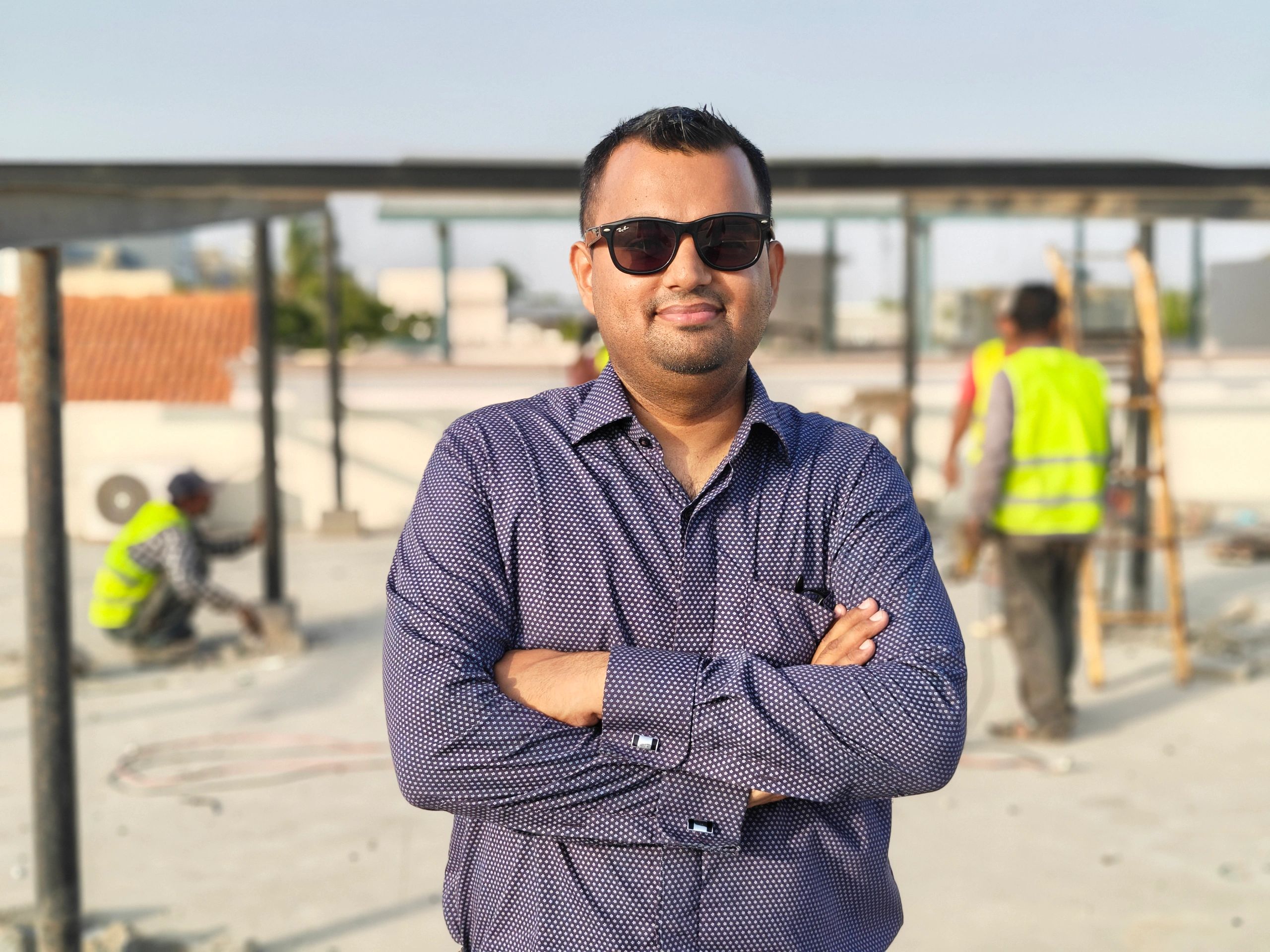 Man in sunglasses smiling with construction workers in background.