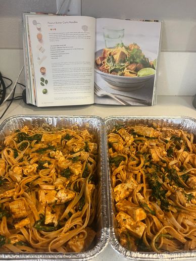 Prepared aluminum pans of peanut Thai noodles with cookbook in background.