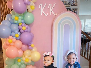 Two toddlers posing at a colorful birthday backdrop with balloons and rainbow decor.