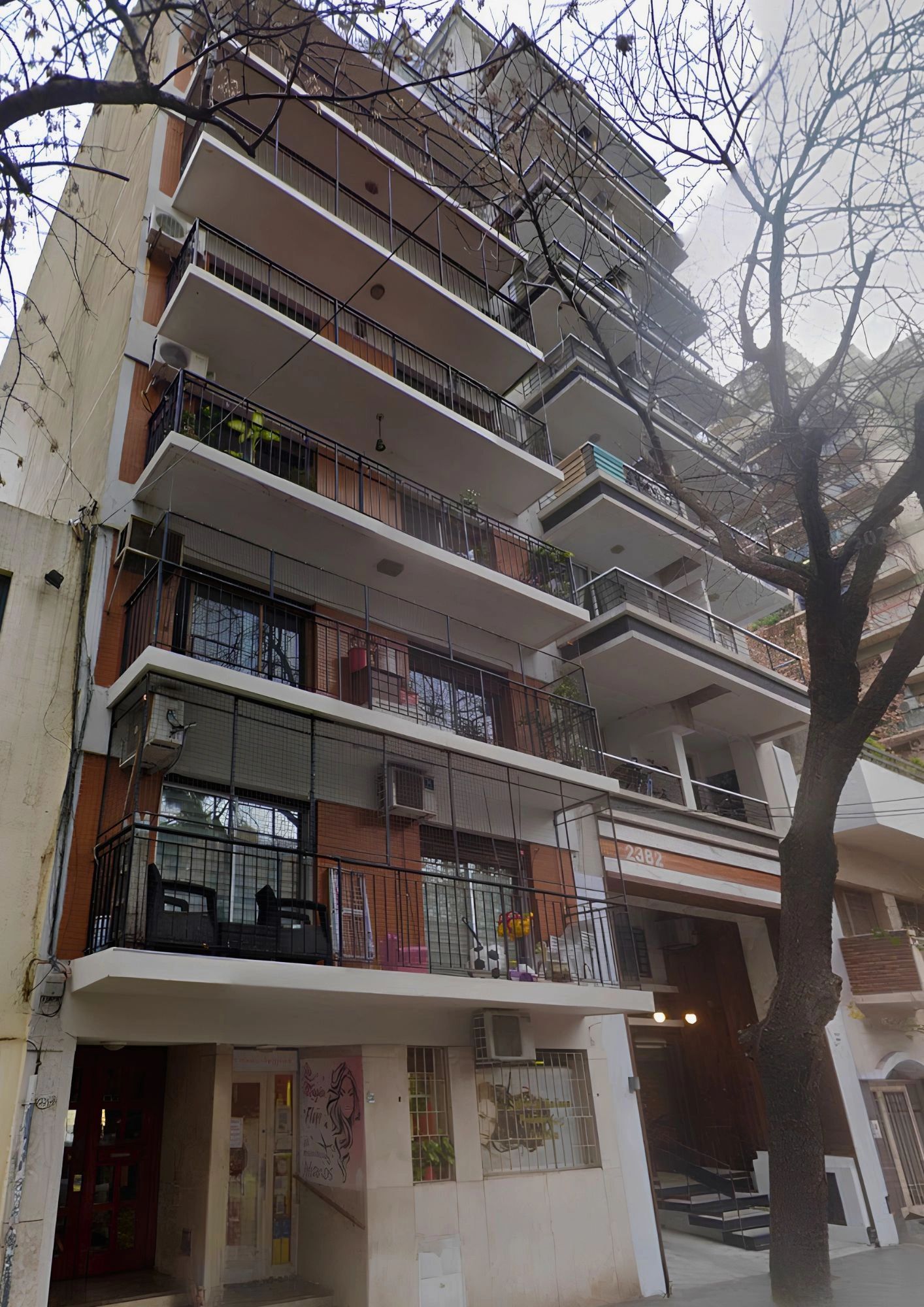 Edificio de departamentos con balcones y árbol frente.