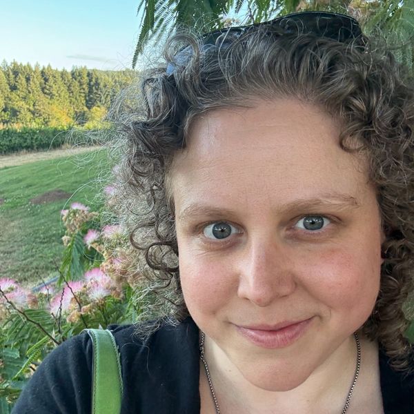 Woman with curly hair smiling outdoors with greenery and flowers in the background.