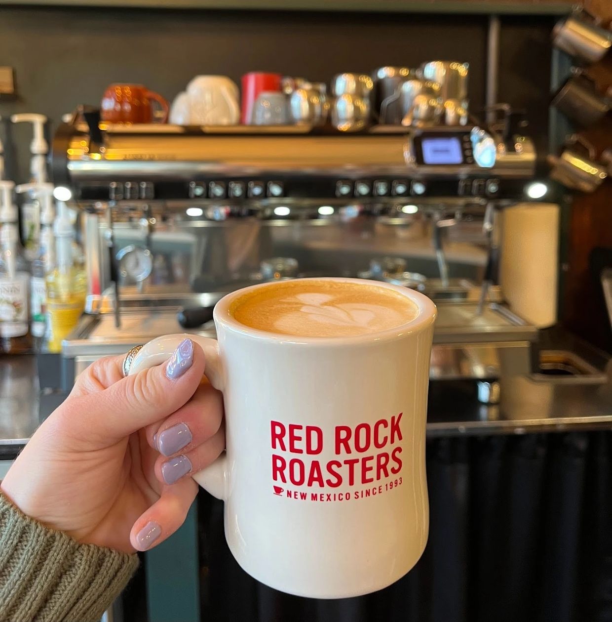 Hand holding a Red Rock Roasters coffee mug with latte art in a café.