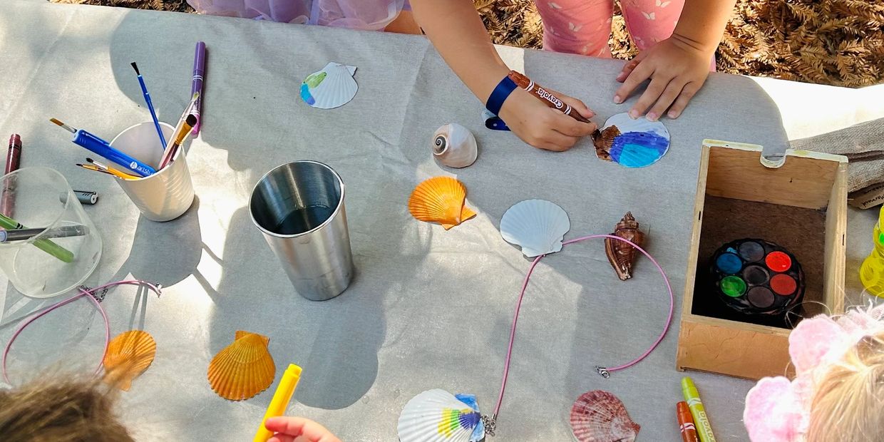 Children decorating seashells with markers and paints on a table outdoors.
