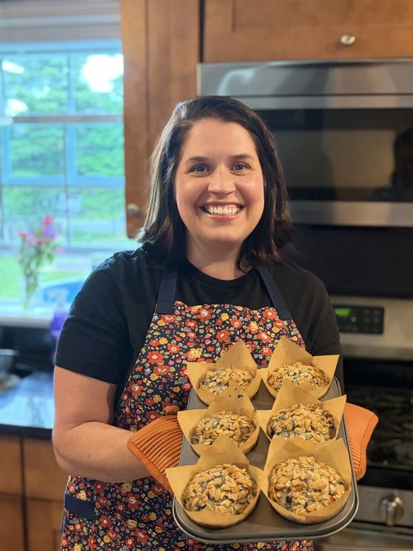 Smiling woman holding a tray of muffins in a kitchen.