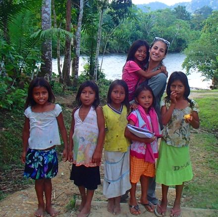 Dr Paula Tallman with various female children in colorful clothing on a riverbank in Peru amazon