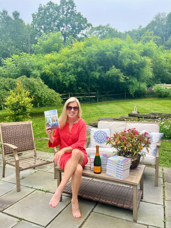Woman in red dress holding a book outdoors with multiple copies on a table.