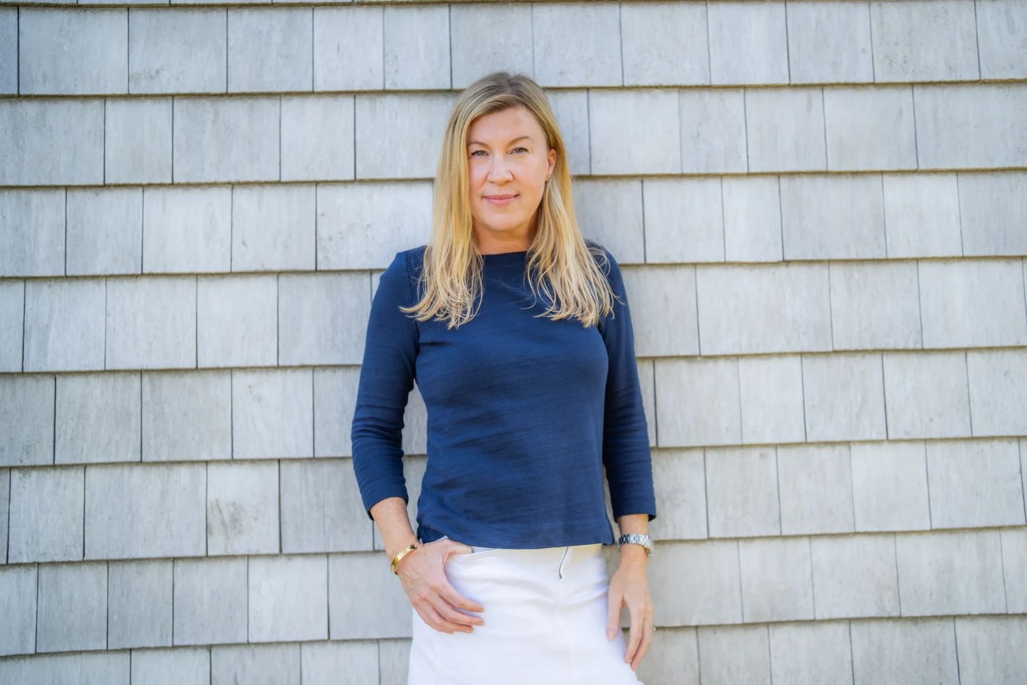 Blonde woman in navy top and white skirt standing against a tiled wall.