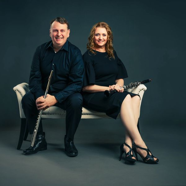 Two musicians pose with flute and clarinet on a bench.