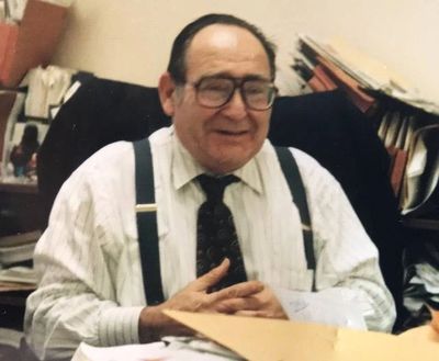 Elderly man in glasses and suspenders smiling at a desk.