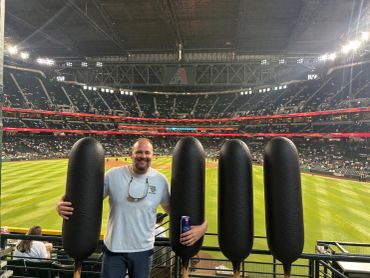 Man posing at baseball stadium with large inflatable sausages.