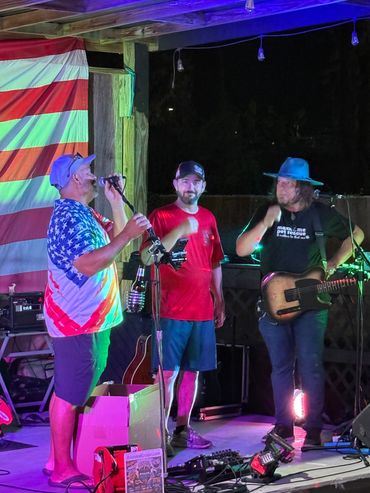 Three men on stage with musical instruments and American flag backdrop.