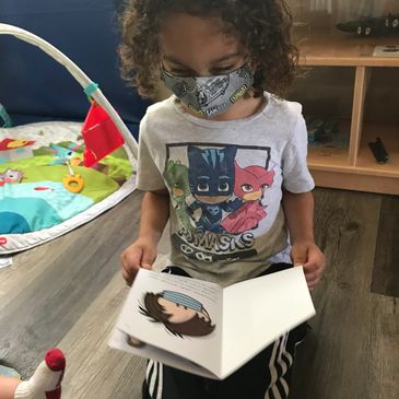 Child with curly hair wearing a cartoon face mask reading a book.