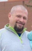 A smiling man with a shaved head and beard in a light gray top.