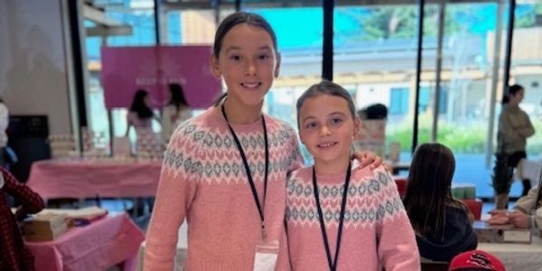 Two girls wearing matching pink sweaters with patterned yokes smile indoors.