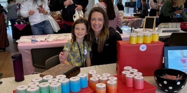 A woman and girl smiling behind a table with colorful candles at a market.