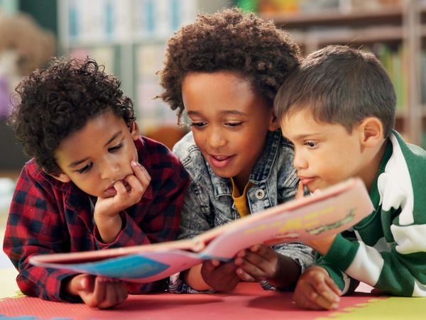 A group of young children are lying on their stomachs on a colorful mat, looking at a picture book.
