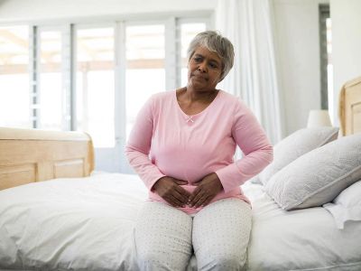 Older woman sitting on bed, holding her lower abdomen in discomfort.
