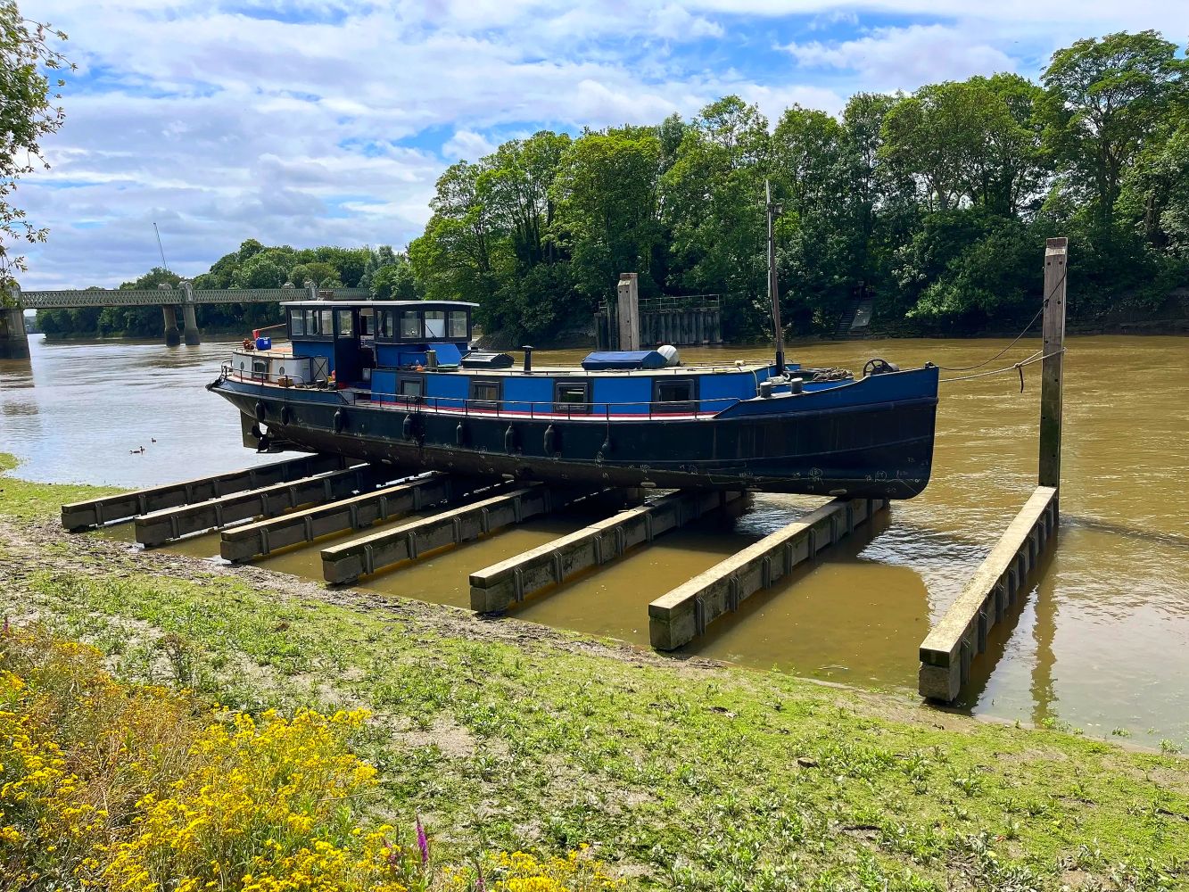 Steel Dutch bargedocked on wooden beams by kew riverbank under a partly cloudy sky.