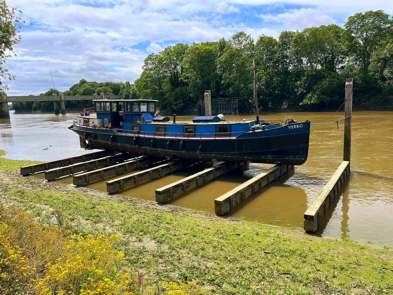 Steel Dutch barge named Verbo docked on wooden beams by kew riverbank under a partly cloudy sky.