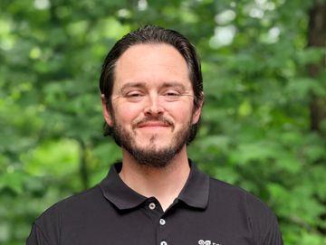Smiling man in a black polo shirt standing outdoors with green trees in the background.