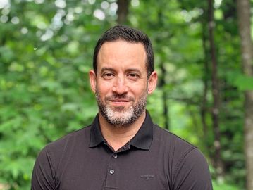 Man with short dark hair and beard wearing a black polo shirt outdoors.