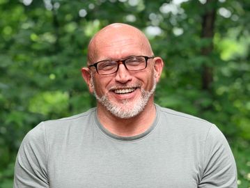 Smiling bald man with glasses outdoors in a green setting.