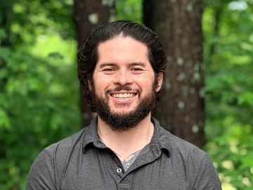 Smiling man with beard standing outdoors in front of trees.