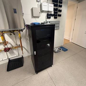Black server rack cabinet in a utility room with pipes and electrical panels.