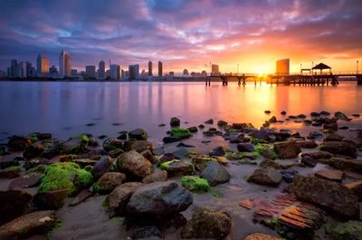 Sunset over a city skyline with rocky shore in the foreground.