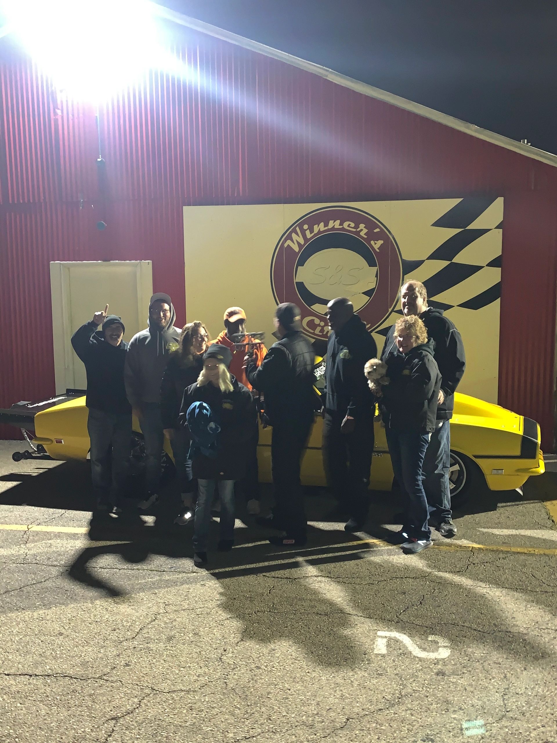Group of people posing at night in front of a race car and sign.