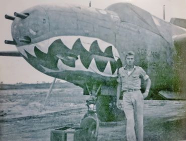 A soldier stands beside a WWII fighter plane with shark mouth nose art.