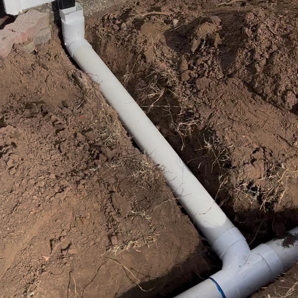 White PVC drainage pipe installed in a dirt trench beside a building.