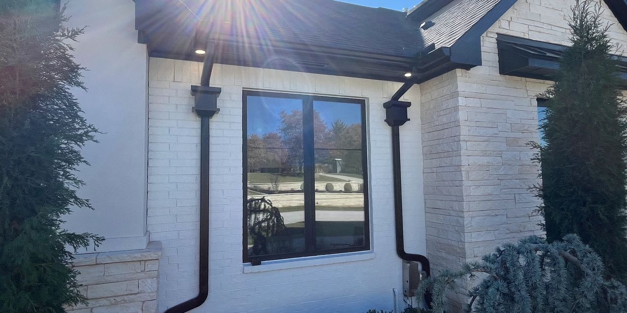 Sunlight shines on a white brick house with a black-framed window and dark gutters.