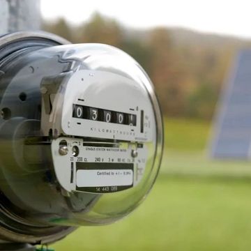 Electric meter with solar panels in the background outdoors.