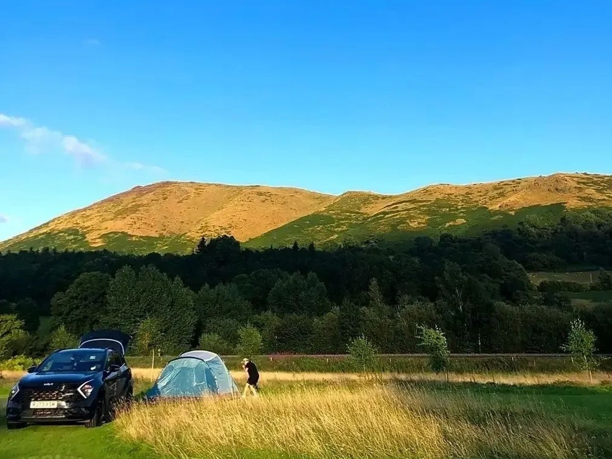 A car and tent set up in a grassy field near forested hills under a clear blue sky.