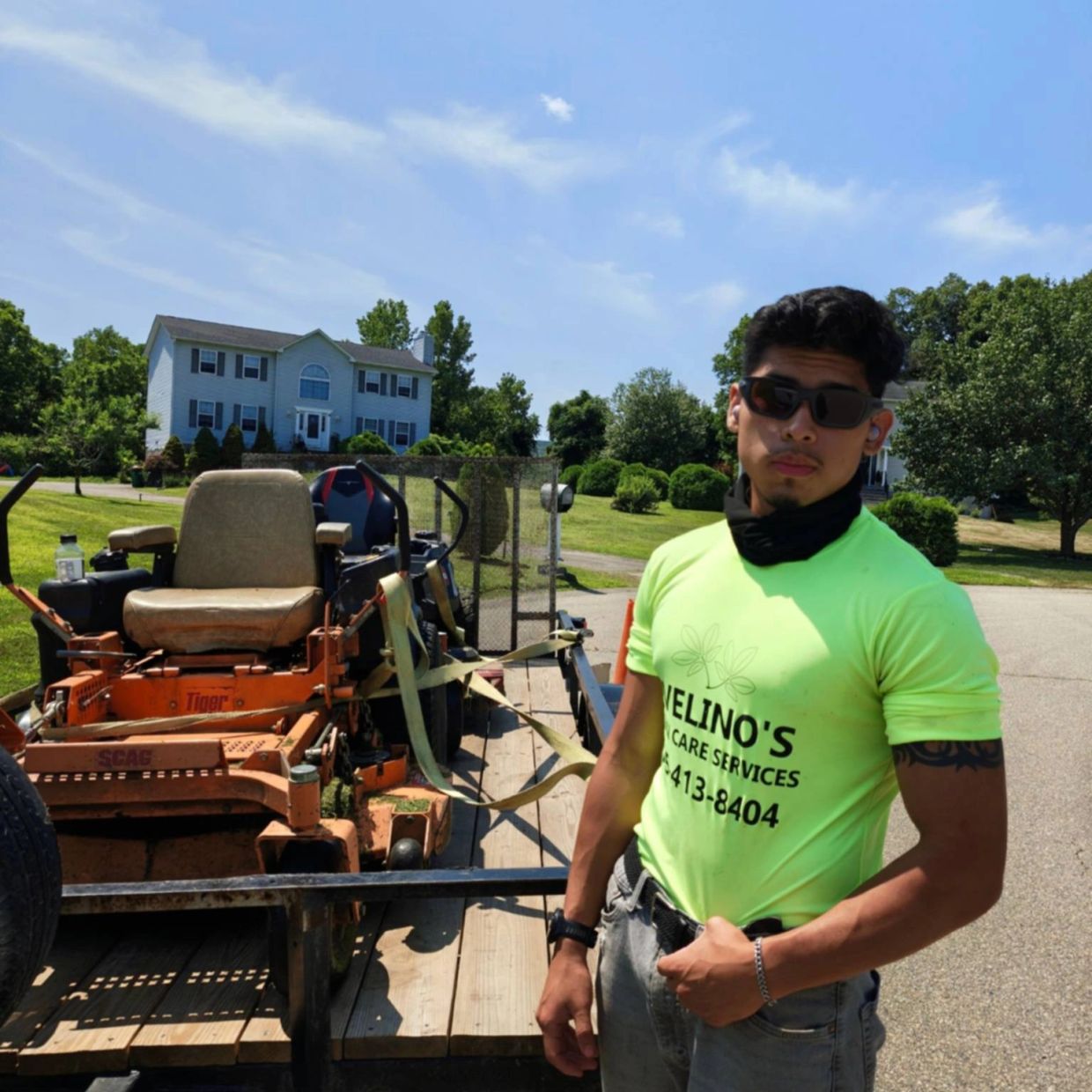 Man in neon shirt standing by lawnmower on trailer in a suburban neighborhood.
