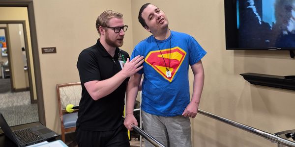 A therapist assists a young man in a Superman shirt during physical therapy.