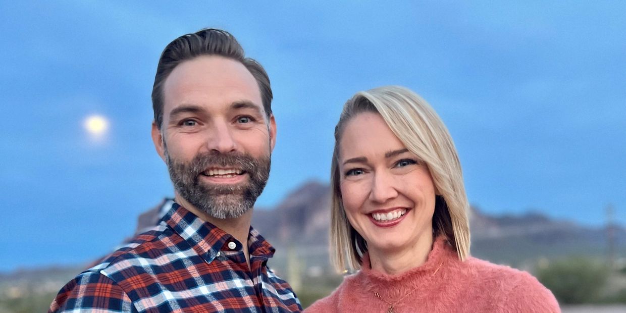 A smiling man and woman posing outdoors with a mountain and moon in the background.