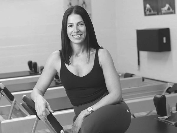 A smiling woman in workout attire sits on Pilates equipment in a fitness studio.