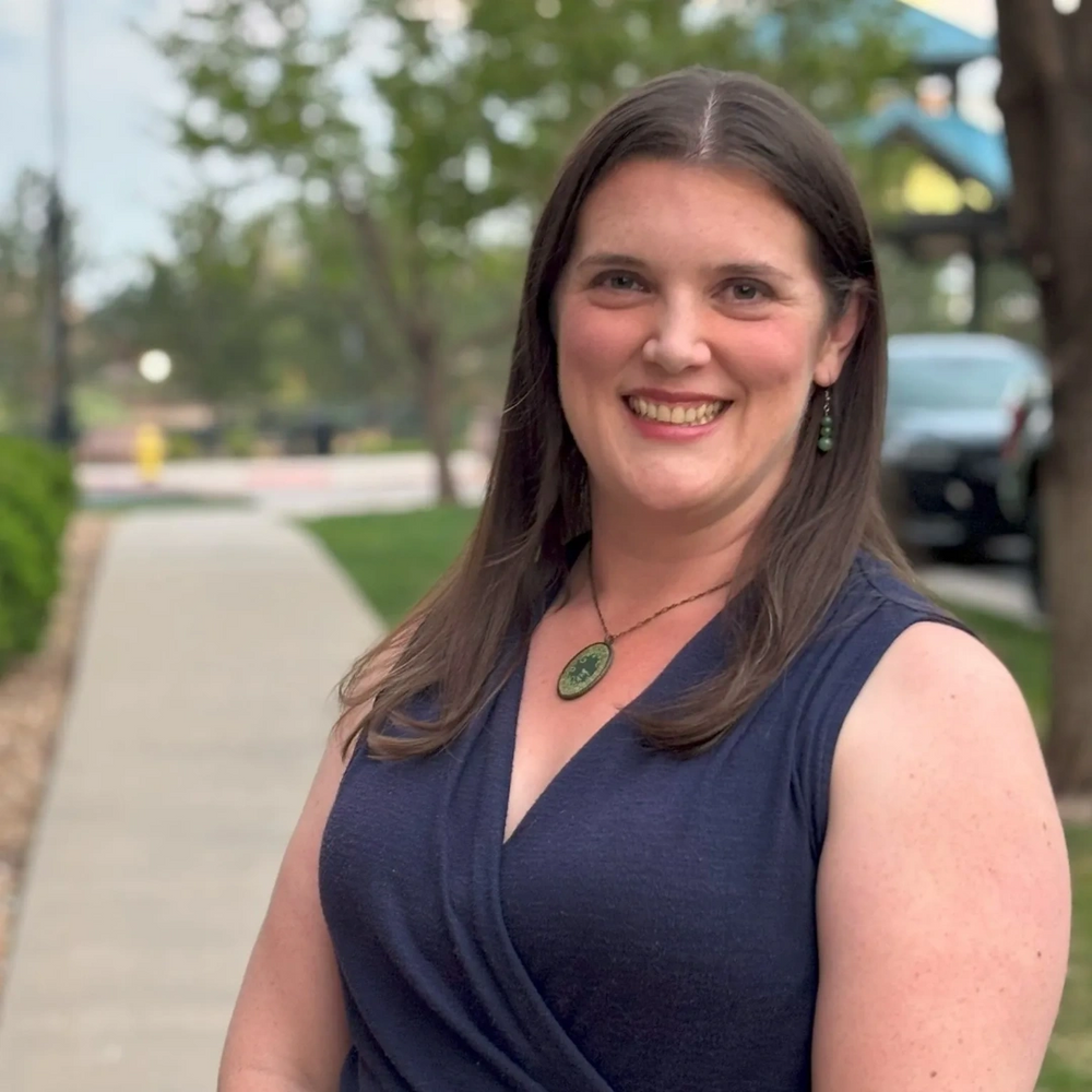 Portrait of Dr. McGill standing on a tree-lined street in a dark blue blouse and green jewelry.