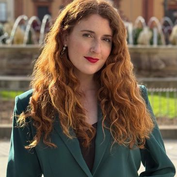 A woman with long red hair wearing a green blazer stands outdoors.