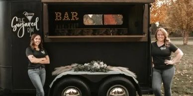 Two women stand beside a decorated mobile bar trailer outdoors.
