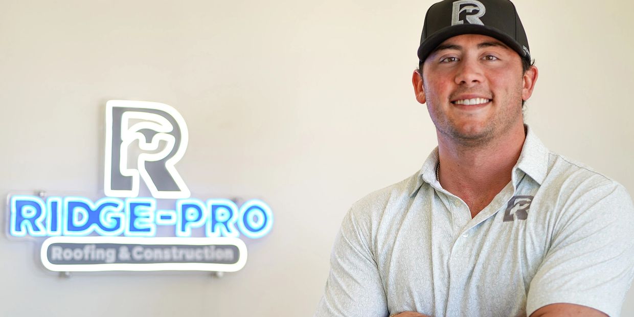 A man standing confidently in front of a Ridge-Pro Roofing &amp; Construction sign.