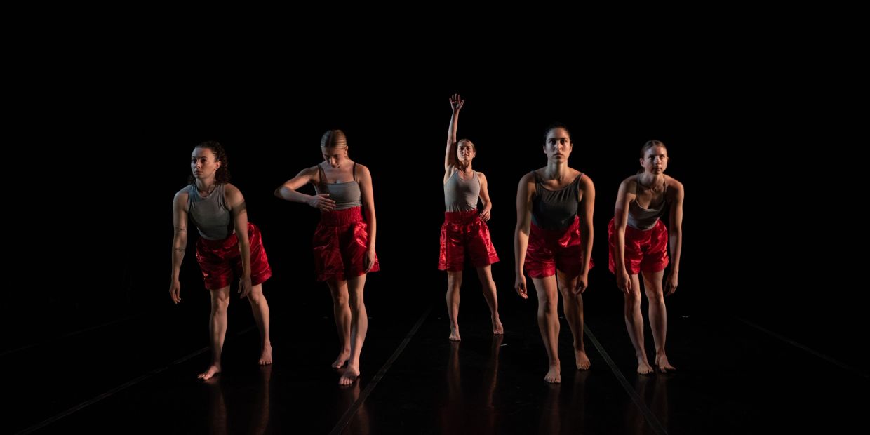 Five dancers in red shorts performing on a dark stage.