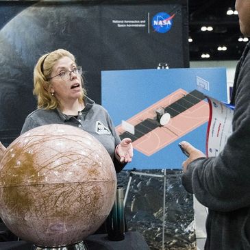 Ocean speaking at an event wearing a gray mission shirt with a globe of the moon Europa.