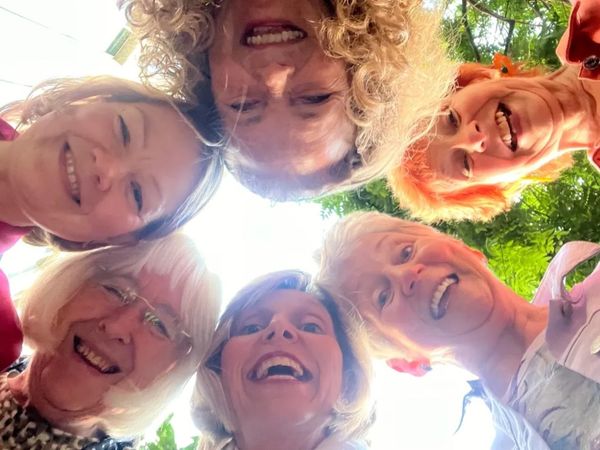 Six members of Vocal Infinity stand in a circle outdoors, looking down toward the camera with trees 