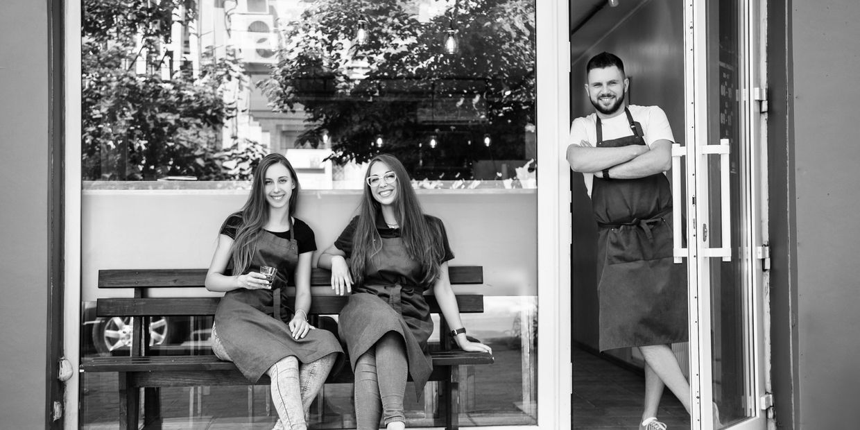 Three smiling cafe staff in blue aprons outside their establishment.